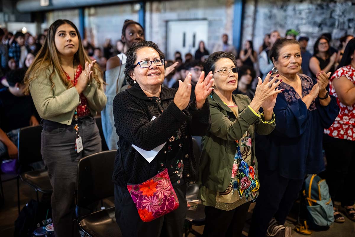 A group of women stands and claps during a Castro event at Metropolitan State University of Denver. The attendees are smiling, showing their appreciation, with others in the background also standing and clapping."